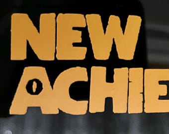 May include: A close-up of a dark surface with the words "NEW ACHIEVEMENT!" in a bold, orange font. The text is centered and takes up most of the frame. The exclamation point is prominent. The background is blurred.