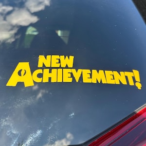 Könnte beinhalten: Gelber Vinyl-Aufkleber mit den Worten "NEW ACHIEVEMENT!" auf einer Autoscheibe. Der Text ist in einer fetten, serifenlosen Schriftart gehalten. Der Buchstabe "A" in "ACHIEVEMENT" hat ein kreisförmiges Design. Der Aufkleber ist auf einer dunklen Oberfläche angebracht.