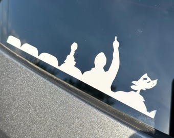 May include: White vinyl decal featuring a crowd silhouette with a person pointing upwards and a cartoon character. The decal is applied to a car window, with the vehicle's gray body visible.