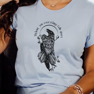 May include: Light blue t-shirt featuring a detailed black raven illustration with the text "Without you everything falls apart" in a circular design. The raven is perched on a branch with small leaves. The shirt has short sleeves.