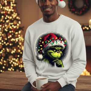 Peut inclure: Sweat-shirt blanc avec un motif du Grinch portant un bonnet de Père Noël orné de guirlandes lumineuses. Le Grinch est vert et affiche une expression espiègle. La personne porte un bonnet de Père Noël et tient une tasse blanche.