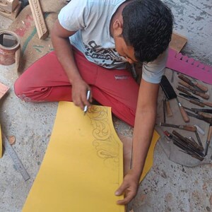 Puede incluir: Una persona trabaja en una pieza de madera amarilla, esbozando un dise&ntilde;o decorativo con un bol&iacute;grafo. Varias herramientas de tallado y elementos de madera est&aacute;n dispersos, lo que sugiere un proyecto de carpinter&iacute;a o artesan&iacute;a. La persona lleva una camiseta azul claro y pantalones rojos.