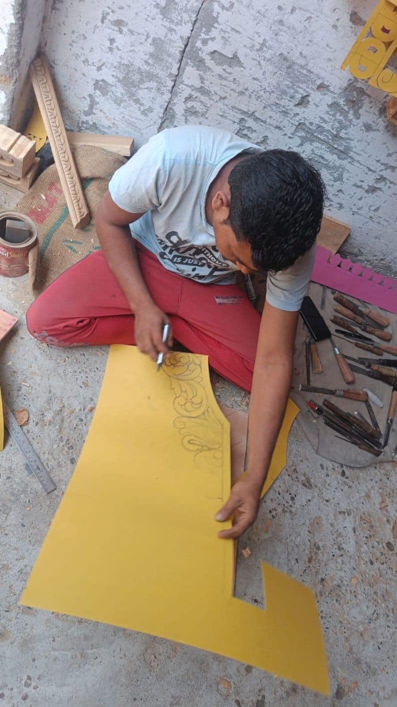Puede incluir: Una persona trabaja en una pieza de madera amarilla, esbozando un dise&ntilde;o decorativo con un bol&iacute;grafo. Varias herramientas de tallado y elementos de madera est&aacute;n dispersos, lo que sugiere un proyecto de carpinter&iacute;a o artesan&iacute;a. La persona lleva una camiseta azul claro y pantalones rojos.