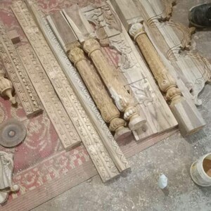 Puede incluir: Elementos arquitect&oacute;nicos de madera tallada, incluyendo columnas y paneles decorativos, dispuestos sobre una alfombra y suelo de hormig&oacute;n. Varias herramientas de carpinter&iacute;a est&aacute;n dispersas cerca, sugiriendo un taller. Las piezas est&aacute;n sin terminar.