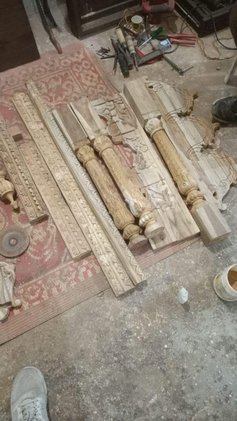 Puede incluir: Elementos arquitect&oacute;nicos de madera tallada, incluyendo columnas y paneles decorativos, dispuestos sobre una alfombra y suelo de hormig&oacute;n. Varias herramientas de carpinter&iacute;a est&aacute;n dispersas cerca, sugiriendo un taller. Las piezas est&aacute;n sin terminar.