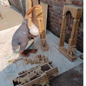 Puede incluir: Un artesano trabajando en muebles de madera tallada intrincadamente. La imagen muestra varios componentes, incluyendo pilares, paneles decorativos y una estructura parcialmente ensamblada, todo en un tono de madera natural. El fondo presenta una pared de ladrillo.