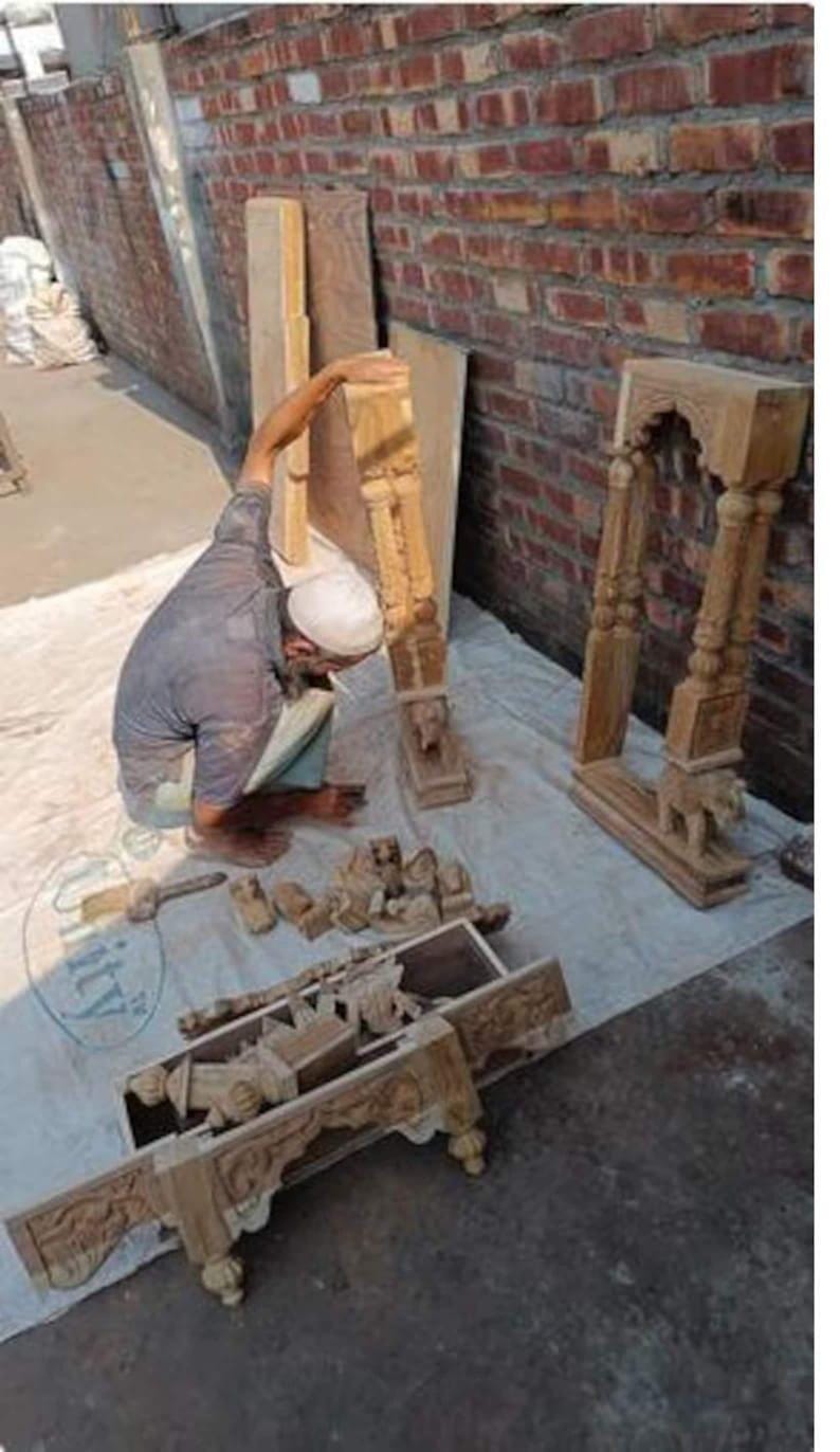 Puede incluir: Un artesano trabajando en muebles de madera tallada intrincadamente. La imagen muestra varios componentes, incluyendo pilares, paneles decorativos y una estructura parcialmente ensamblada, todo en un tono de madera natural. El fondo presenta una pared de ladrillo.