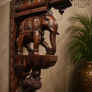 Puede incluir: Un soporte de pared de madera tallada en color marrón oscuro que presenta una figura de elefante detallada en el centro y una figura de ave en la base. La estructura incluye intrincados patrones de volutas a lo largo del soporte vertical.