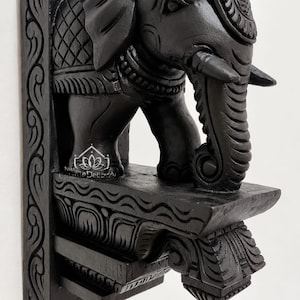 Puede incluir: Un soporte de pared de madera negra con una figura de elefante tallada en la parte superior. El soporte tiene patrones intrincados a lo largo del lateral y la base. Una pequeña lámpara de aceite de latón en forma de estrella cuelga.