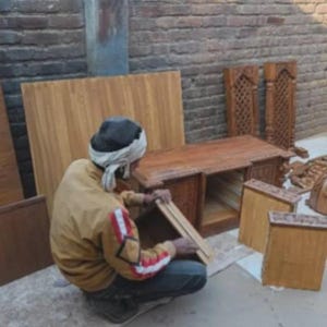 Puede incluir: Una persona ensamblando muebles de madera al aire libre. La escena incluye un escritorio parcialmente construido, cajones y otros componentes de madera. La madera tiene un tono c&aacute;lido y natural, con una pared de ladrillo en el fondo.