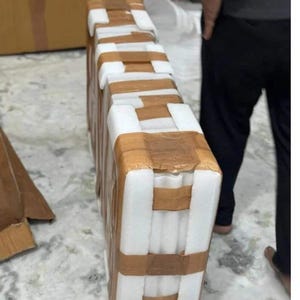 Puede incluir: Un paquete rectangular envuelto en espuma blanca y asegurado con cinta adhesiva marr&oacute;n. La cinta forma bandas horizontales y verticales. El fondo muestra una persona y otras cajas.