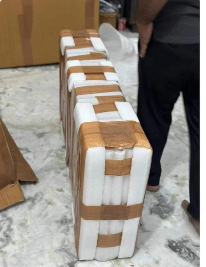 Puede incluir: Un paquete rectangular envuelto en espuma blanca y asegurado con cinta adhesiva marr&oacute;n. La cinta forma bandas horizontales y verticales. El fondo muestra una persona y otras cajas.