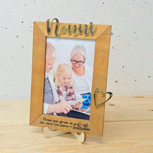 Può includere: Cornice in legno con una foto di tre persone. La parola "Nonni" è in oro nella parte superiore. Un cuore d'oro è sul lato destro. La cornice ha un supporto in legno e una citazione in italiano.