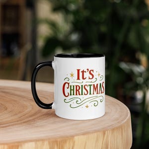 May include: White ceramic mug with a black handle and interior. The mug features the text "It's Christmas" in red and green, with decorative swirls and stars. The mug is sitting on a wooden surface.