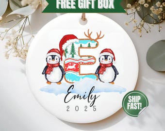 May include: A white ceramic Christmas ornament featuring two penguins wearing Santa hats and red scarves. The letter "E" is decorated with a Christmas theme. The ornament is personalized with the name "Emily" and the year "2025".
