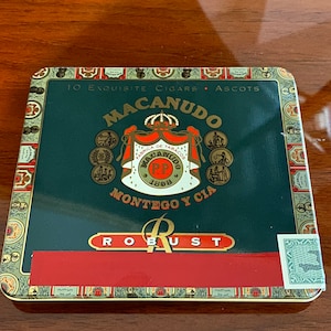 May include: A green and gold tin box with a red band and the words "10 Exquisite Cigars Ascots Macanudo Fabrica de Tabacos Macanudo S.P.P. 1888 Montego y Cia Robust" printed on it.