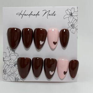 Könnte beinhalten: Ein Set handgefertigter Press-On-Nägel in Braun- und Rosatönen. Einige Nägel haben ein Herzdesign. Auf der Karte steht in Schreibschrift "Handmade Nails". Präsentation auf weißem Hintergrund.