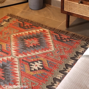 May include: A rectangular area rug with a geometric pattern in shades of red, gray, and beige. The rug has a fringed edge and a repeating diamond and triangle design. The text "CarpetEmpires" is visible in the bottom left corner.