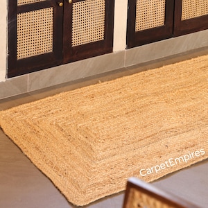 May include: A rectangular jute rug in a natural colour, featuring a woven design. The rug is positioned on a grey floor, with a dark brown cabinet with woven cane detailing in the background. The words "CarpetEmpires" are visible in the bottom right corner.