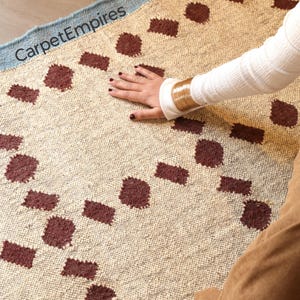 May include: Beige and burgundy patterned rug with a light blue border. The rug features a repeating pattern of burgundy squares and circles. The text "CarpetEmpires" is visible on the rug's border.