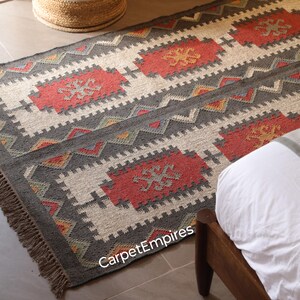 Puede incluir: Una alfombra rectangular con un patrón geométrico. Tiene un borde gris con un diseño en zigzag. El diseño central incluye formas rectangulares rojas y beige con un patrón en forma de cruz. La alfombra tiene un borde con flecos y el texto "CarpetEmpires".