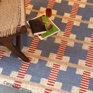 Peut inclure: Tapis à motifs bleus, beiges et rouges avec un motif géométrique. Une chaise en bois avec une assise tissée est à gauche. Un verre de vin et un livre reposent sur le tapis. Le texte "Carpet Empires" est visible.