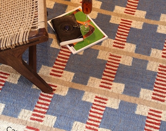 May include: A blue, beige, and red patterned rug with a geometric design. A wooden chair with a woven seat is on the left. A glass of wine and a book rest on the rug. The text "Carpet Empires" is visible.