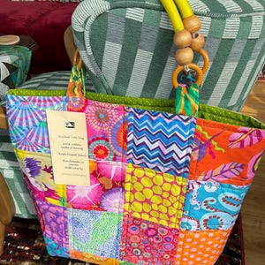 May include: A patchwork tote bag with a yellow handle and wooden beads. The bag is made from a variety of colourful fabrics with floral, geometric, and polka dot patterns. The bag has a green lining and a tag that reads "Quilted Tote bag".