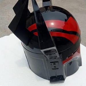 Puede incluir: Un casco negro y rojo con un acabado brillante. El casco presenta una parte superior redondeada con rayas rojas y prominentes acentos angulares negros. El dise&ntilde;o sugiere un tema futurista o de ciencia ficci&oacute;n.