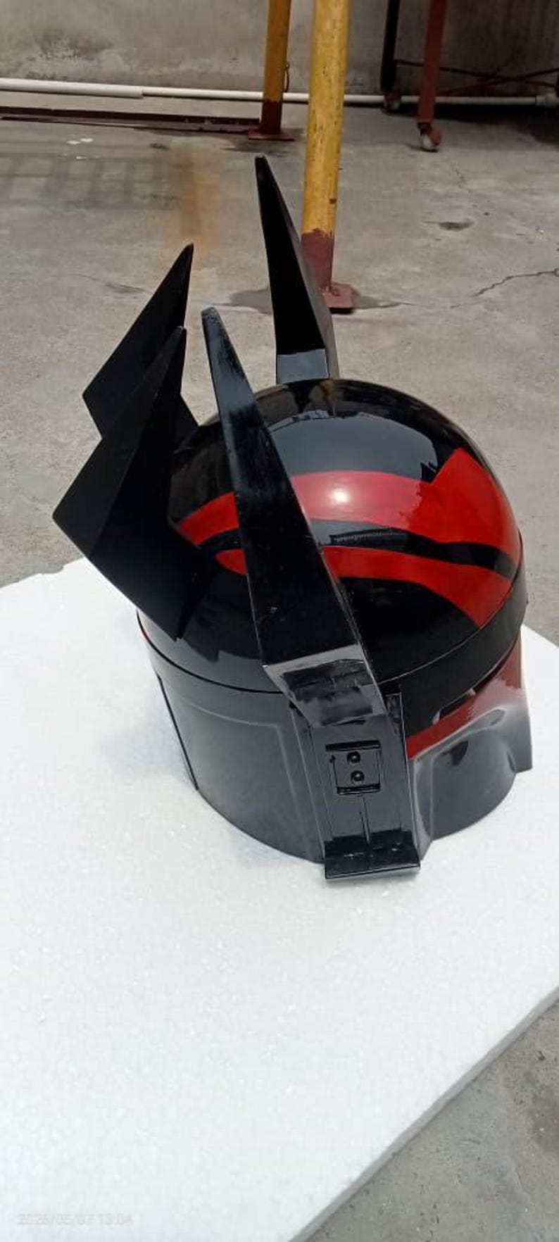 Puede incluir: Un casco negro y rojo con un acabado brillante. El casco presenta una parte superior redondeada con rayas rojas y prominentes acentos angulares negros. El dise&ntilde;o sugiere un tema futurista o de ciencia ficci&oacute;n.