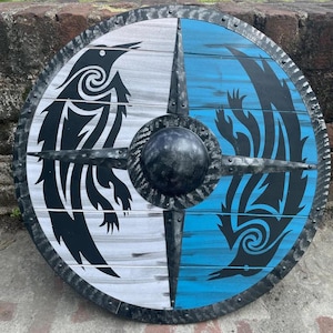 Puede incluir: Un escudo redondo de madera con un borde de metal negro y un centro abultado. El escudo está dividido en cuatro secciones: dos blancas y dos azules. Cada sección de color presenta un diseño negro estilizado de pájaro o cuervo. El escudo tiene una apariencia rústica y artesanal.