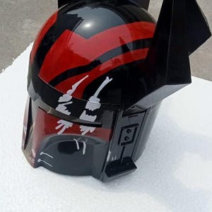 Puede incluir: Un casco negro y rojo con detalles angulares y puntiagudos. El casco presenta un acabado brillante con rayas rojas y detalles gr&aacute;ficos blancos. El dise&ntilde;o sugiere un tema futurista o de ciencia ficci&oacute;n.