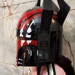 Puede incluir: Un casco negro y rojo con un dise&ntilde;o &uacute;nico, con formas angulares y un acabado brillante. El casco tiene acentos negros prominentes y una franja roja. El dise&ntilde;o incluye detalles gr&aacute;ficos blancos.