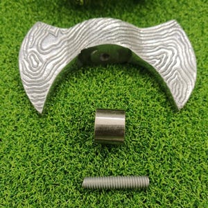 Puede incluir: Un tirador de armario decorativo plateado con un patrón de vetas de madera. También son visibles un pequeño cilindro metálico dorado y un tornillo plateado. Los artículos están dispuestos sobre una superficie verde.