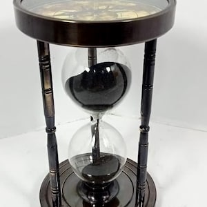 Può includere: Clessidra in bronzo scuro con due globi di vetro trasparente riempiti di sabbia nera. La clessidra ha una base e una parte superiore rotonde ed è sostenuta da quattro pilastri in bronzo scuro. Altezza circa 20 cm.