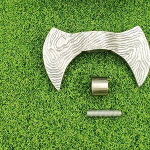 Puede incluir: Una cabeza de hacha de doble filo desmontada con un patrón texturizado de vetas de madera. La cabeza del hacha es plateada y se encuentra sobre una superficie verde, junto con un cilindro de metal plateado y un perno roscado.