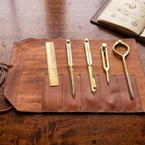 Puede incluir: Colección de herramientas de dibujo antiguas, incluyendo una regla, compases y divisores, exhibidas en un rollo de herramientas de cuero marrón. Un libro abierto con diagramas, una pluma y un mapa enrollado sugieren un tema de artesanía histórica.