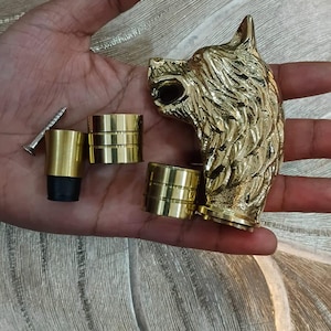 Può includere: Una scultura a forma di testa di lupo color oro con dettagli intricati, insieme a diversi componenti cilindrici color ottone e una vite. La scultura è probabilmente un elemento decorativo o parte di un oggetto più grande.