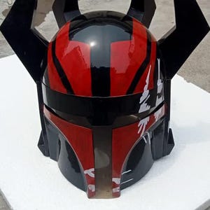 Puede incluir: Un casco negro y rojo con un acabado brillante. El casco presenta un dise&ntilde;o distintivo con formas angulares y cuatro puntas negras prominentes. La parte delantera del casco tiene texto blanco. El casco se muestra sobre una superficie blanca.