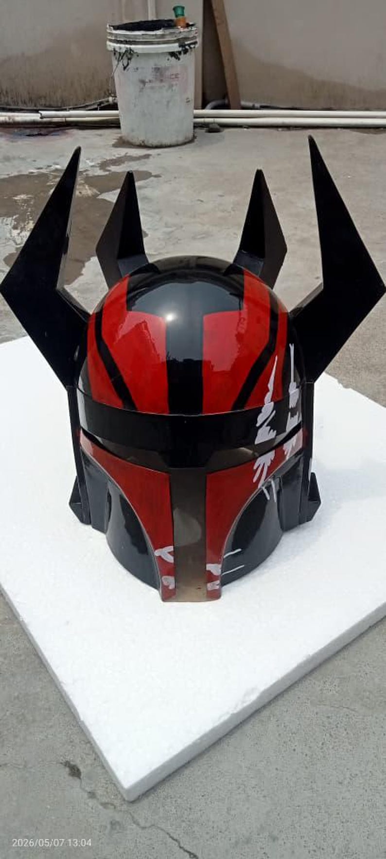 Puede incluir: Un casco negro y rojo con un acabado brillante. El casco presenta un dise&ntilde;o distintivo con formas angulares y cuatro puntas negras prominentes. La parte delantera del casco tiene texto blanco. El casco se muestra sobre una superficie blanca.