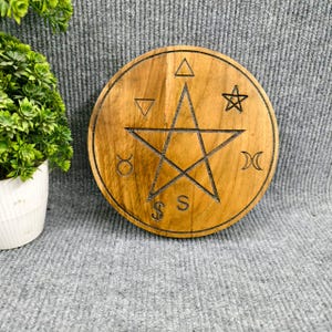 May include: A circular wooden plaque featuring a pentagram design, with etched symbols around the edge. The wood has a warm, natural colour. The item is likely used for spiritual or decorative purposes, with a diameter of approximately 20 cm.