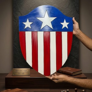 May include: A handcrafted Captain America shield tribute, circa 2024. The shield features a blue top with three white stars and red and white stripes. The shield is held up by two hands.