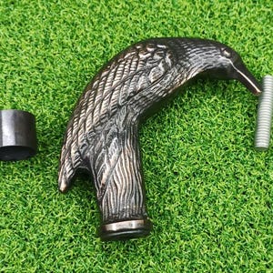 Puede incluir: Un mango de bastón con cabeza de águila de color bronce detallado, con una férula a juego y un tornillo plateado. La cabeza del águila presenta intrincados detalles de plumas. Los artículos están sobre un fondo verde.