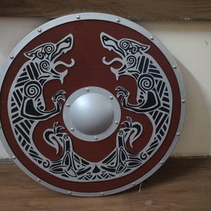 May include: A round, wooden shield with a dark red center and a silver rim. The shield features a symmetrical design of two wolf-like creatures in black and white, with a silver boss in the center. Decorative and functional.