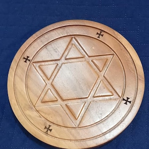May include: A round, wooden decorative item featuring a carved Star of David within a circle. Four cross-like symbols are evenly spaced around the outer edge. The item is made of light-colored wood and sits on a blue surface.