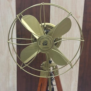 Puede incluir: Un ventilador de latón de estilo vintage con cuatro aspas, encerrado en una jaula protectora. El ventilador está montado sobre un trípode de madera. El centro del ventilador dice "MADE FOR ROYAL NAVY LONDON".