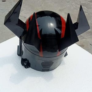 Puede incluir: Casco negro con detalles rojos y protuberancias angulares. El casco tiene un acabado brillante y una ranura de ventilaci&oacute;n en la parte inferior. Est&aacute; sobre una superficie blanca.