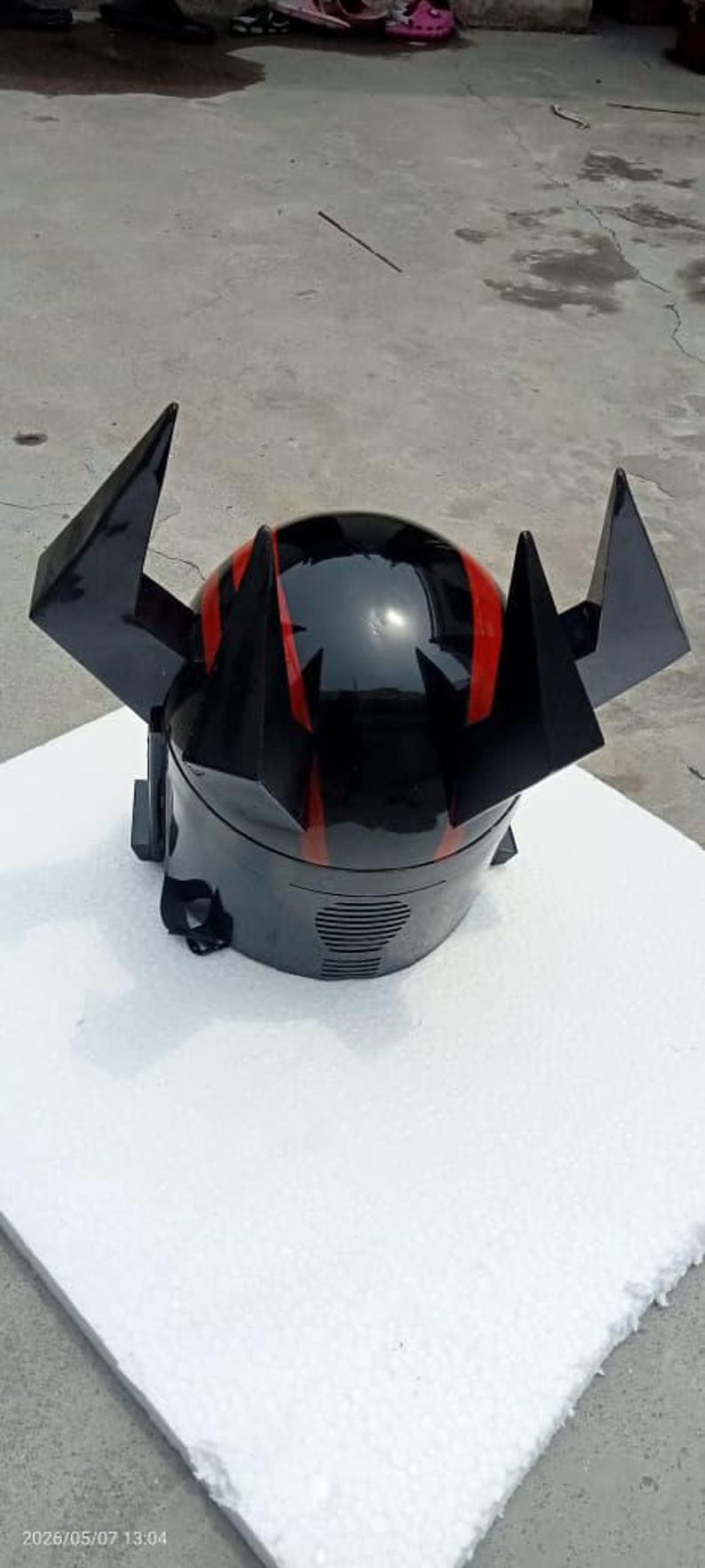 Puede incluir: Casco negro con detalles rojos y protuberancias angulares. El casco tiene un acabado brillante y una ranura de ventilaci&oacute;n en la parte inferior. Est&aacute; sobre una superficie blanca.