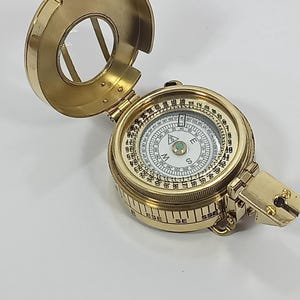 May include: A vintage brass compass with a hinged lid, open to reveal the compass dial. The compass has a circular design with directional markings and a needle. The lid is open, showing the glass cover. The compass is on a white surface.
