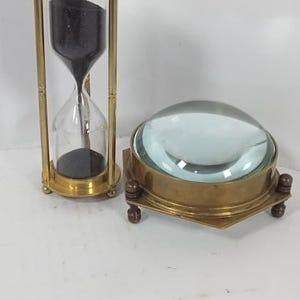 Op de afbeelding: Een zandloper met zwart zand en een messing frame staat naast een vergrootglas met messing frame. De zandloper is een klassiek tijdmeetapparaat, terwijl het vergrootglas een vintage bureau-accessoire is.